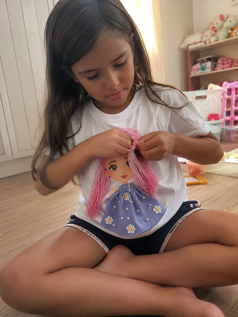 Menina sentada no chão do quarto trançando o cabelo de lã rosa da camiseta Madeixa